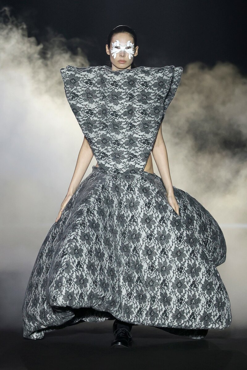 Model on the 080 Barcelona Fashion catwalk wearing a Guillermo Justicia dress with a floral print, large triangular volumes on the bodice and skirt, and gray tones.
