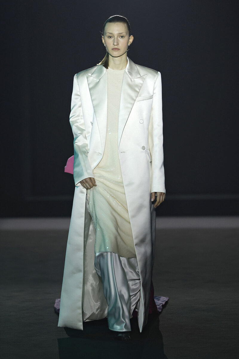 Model parading at 080 Barcelona Fashion in a total white look featuring an extra-long satin coat and sequin dress by designer Juan Vidal.