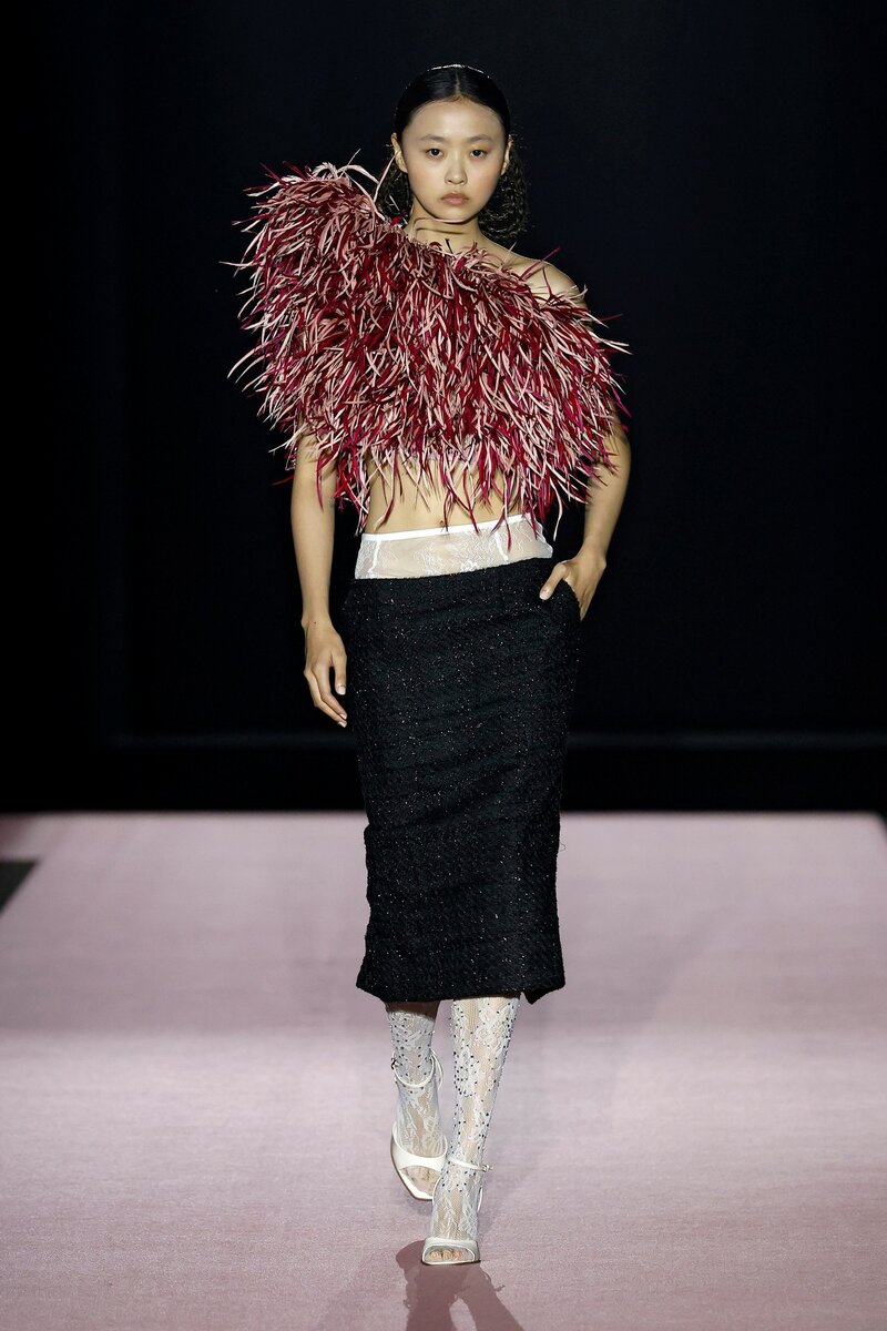 Model showing off a feminine look by COMPTE SPAIN at 080 Barcelona Fashion with an asymmetrical burgundy feather top and a black skirt with a wide white waistband.