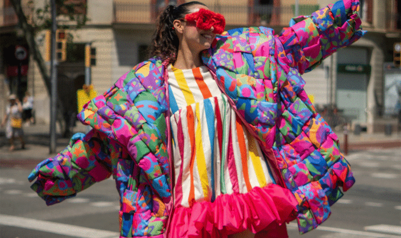 A girl wearing large red glasses and an oversized patchwork coat over a colorful striped dress with a cheerful expression in the middle of the street