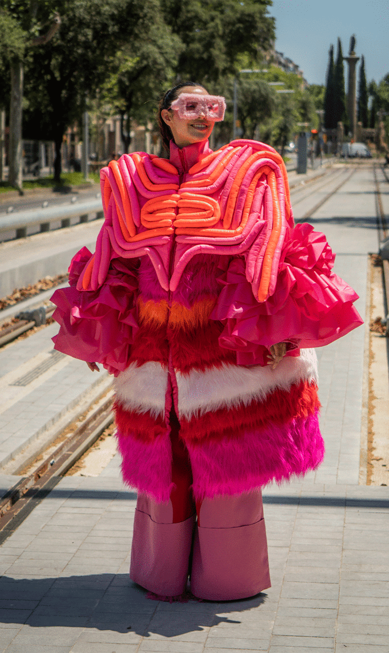 A girl in the middle of the street with large pink glasses and a very large outfit of bright colors in red, pink, orange and white