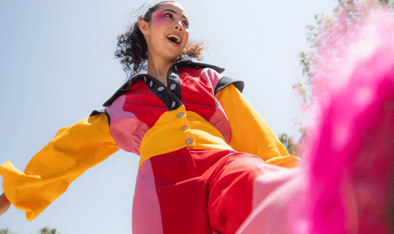 Low angle shot of a cheerful girl kicking her feet in the air. She's wearing a colorblock jumpsuit in shades of red, pink, yellow, and black with a shirt collar.