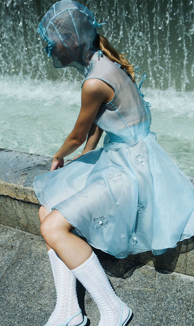 Female model sitting in front of a curtain of water wearing a light blue dress with sheer panels