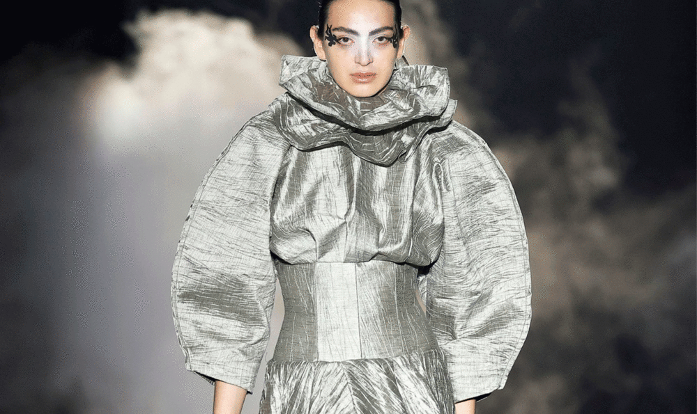 Model wearing a high-neck, ruffled, shiny silver-gray dress with wide, puffed sleeves by IED Barcelona alum Guillermo Justicia at 080 Barcelona Fashion