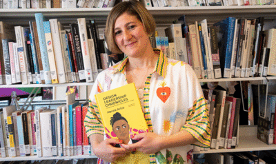 A blonde woman holds a yellow book titled Design Leadership Chronicles in front of a large bookshelf full of books.