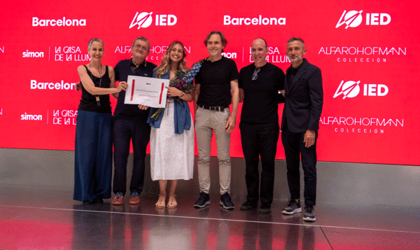 A jury of 5 people presents a diploma to a student on a gray stage with a red screen and IED Barcelona logos in the background.