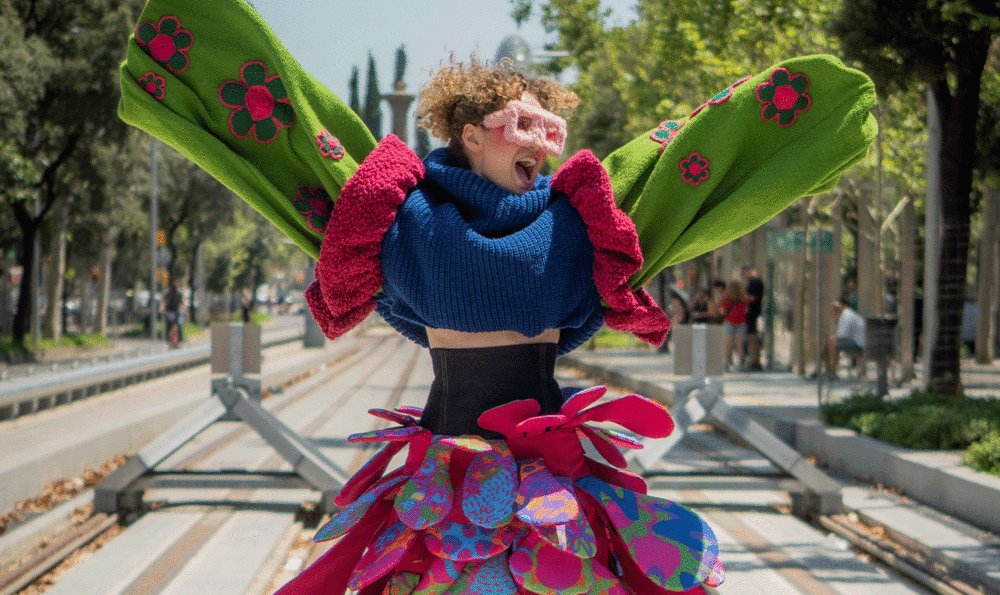 A cheerful young girl with open arms and a happy expression. She's wearing very big sunglasses and very colorful clothes standing in front of the tram tracks.