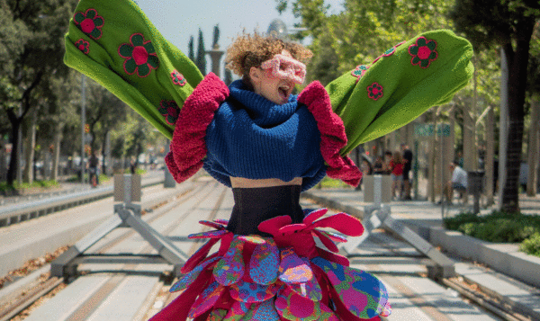 A cheerful young girl with open arms and a happy expression. She's wearing very big sunglasses and very colorful clothes standing in front of the tram tracks.